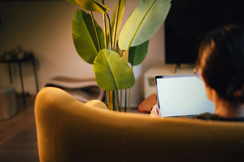 A woman is comfortably using a laptop in a cozy home environment with plants and warm lighting.